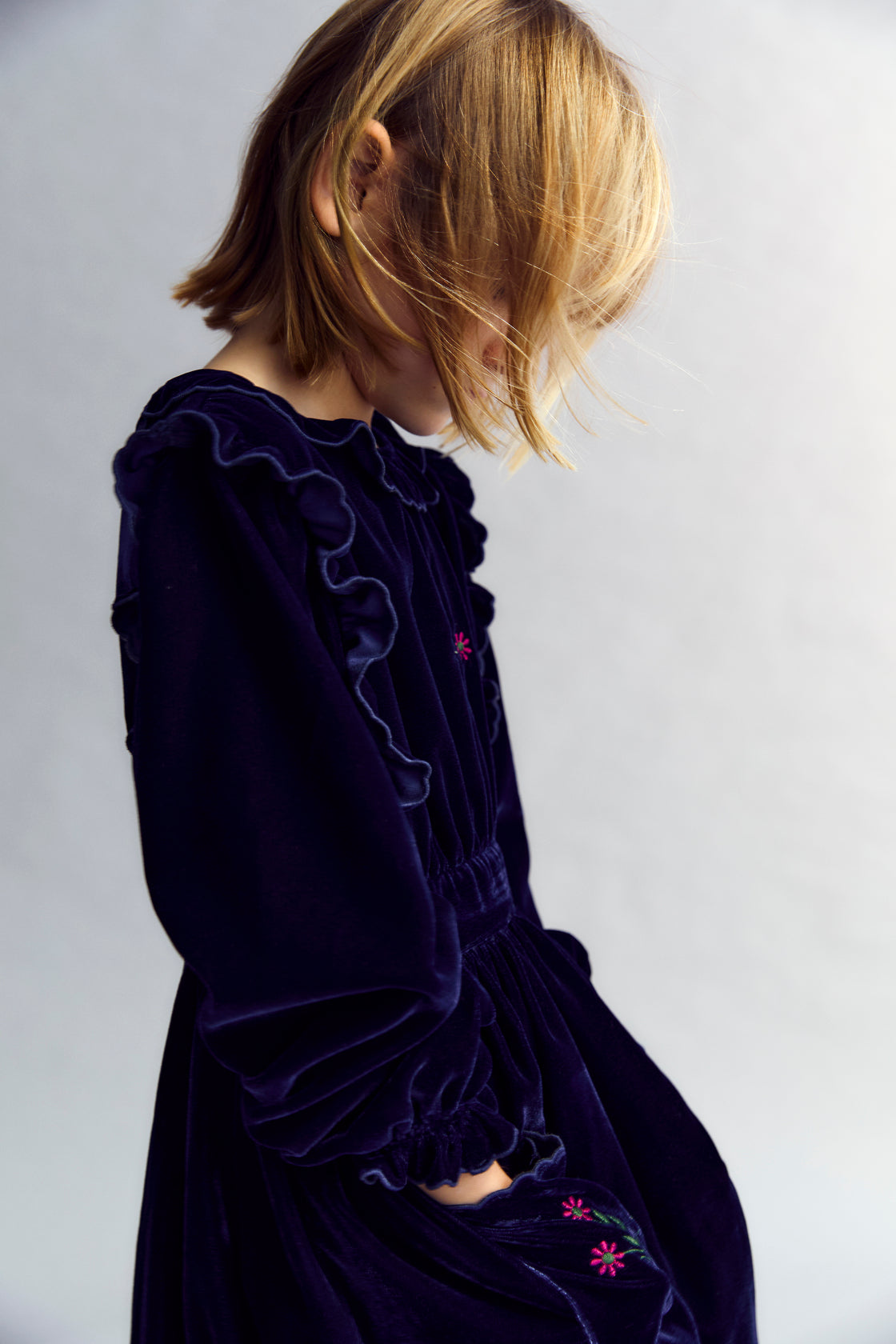 Velour dress with a fitted waist, featuring embroidery detail on the chest and two embroidered pockets on the skirt. It is finished with ruffles at the cuffs and opens at the back with ruffled details. The dress has cotton lining in tone.