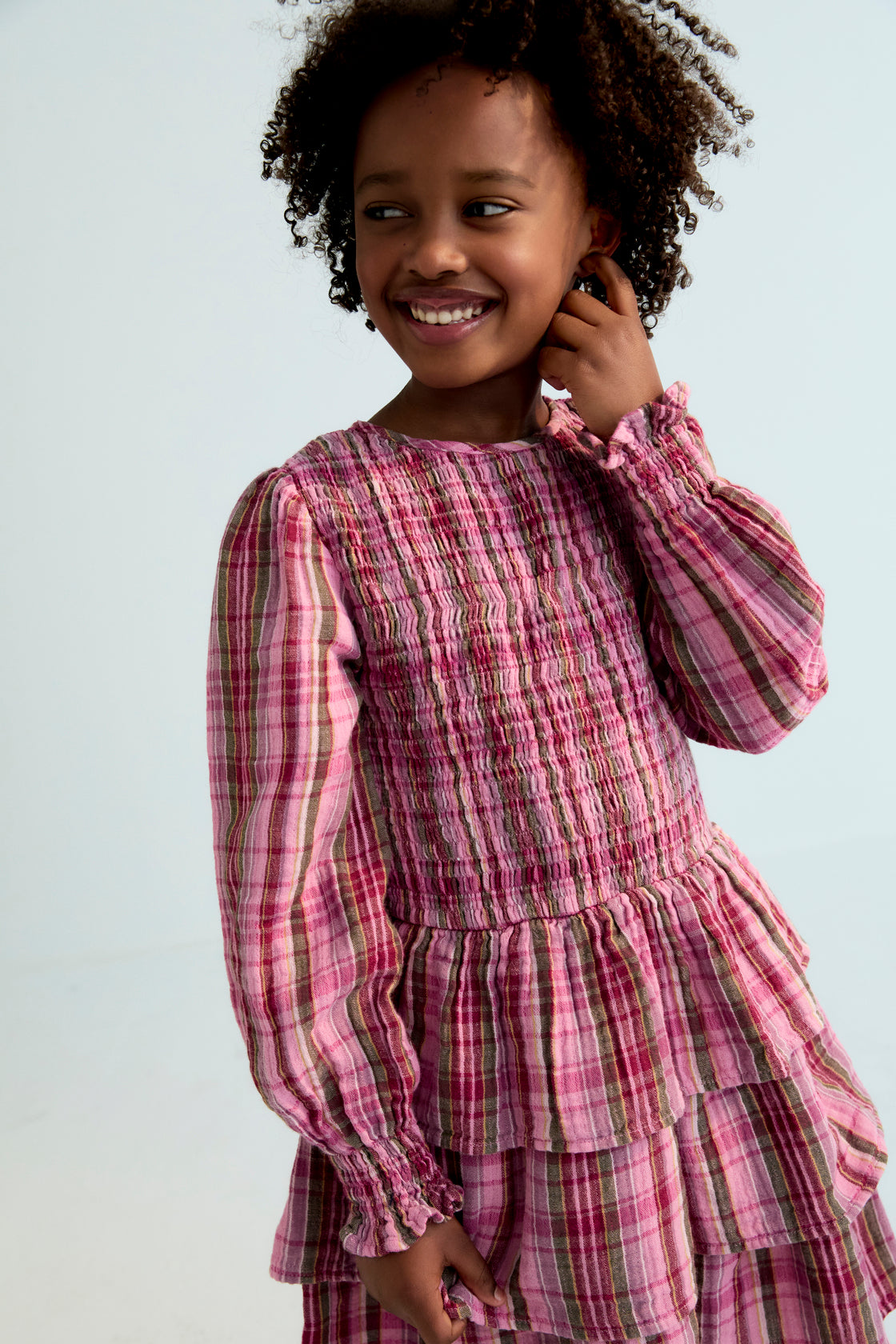 Short checkered double gauze dress with a smocked bodice, elasticated fitted waistband, and three ruffles on the skirt. It features a back opening with a teardrop cut and button closure.
