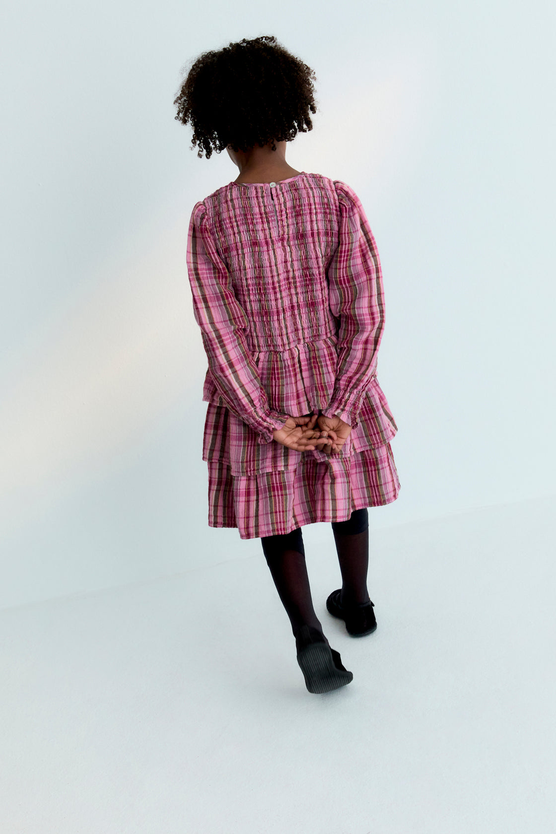 Short checkered double gauze dress with a smocked bodice, elasticated fitted waistband, and three ruffles on the skirt. It features a back opening with a teardrop cut and button closure.