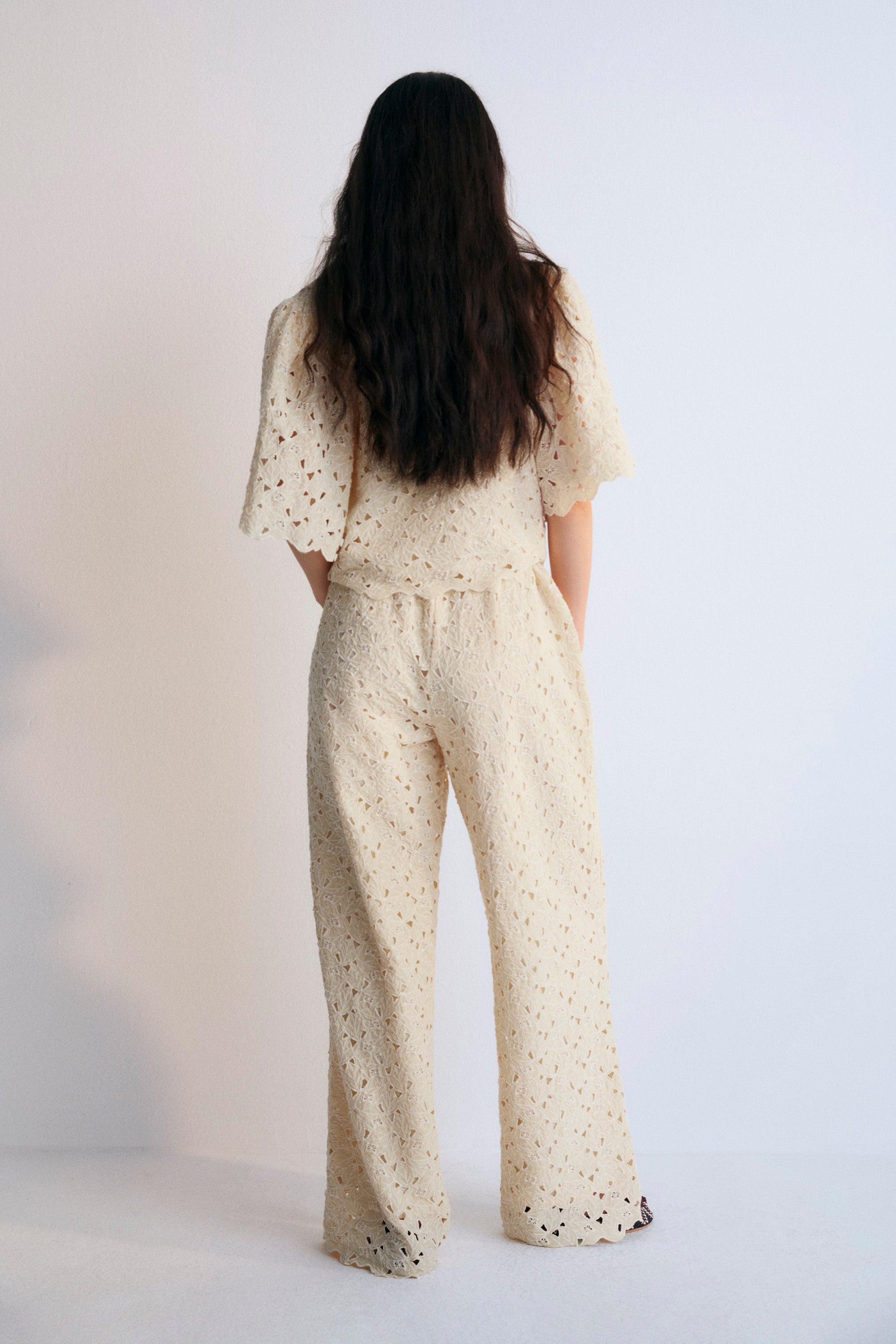 A complete outfit from The New Society's Spring Summer Collection, featuring the Atenea Woman Blouse in embroidered macadamia-colored cotton blend fabric, with gathered sleeves, a V-neckline, and a front closure tied with two bows. Paired with a matching top and pants in the same macadamia tone, creating a cohesive, elegant, and airy look perfect for warmer seasons.