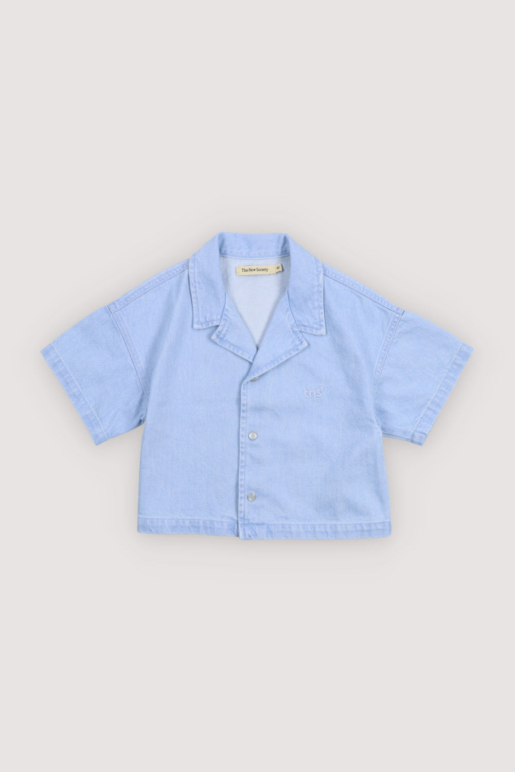 Short sleeve cropped shirt in washed denim, featuring a front snap closure, chest pocket, and TNS heart embroidery.
