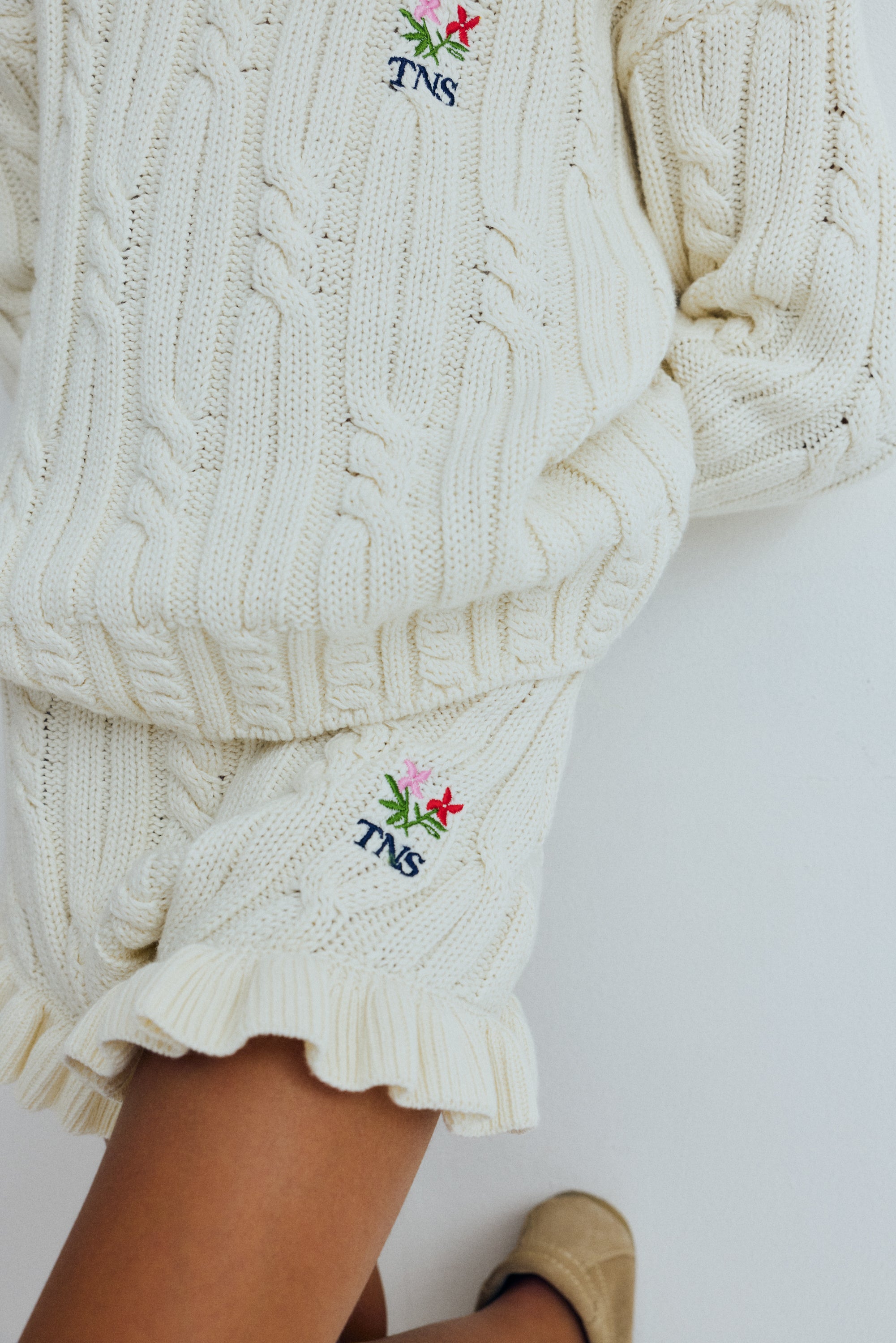 Cable knit sweater with subtle cable details on the collar, cuffs, and hem, featuring a playful floral branded embroidery on the chest.