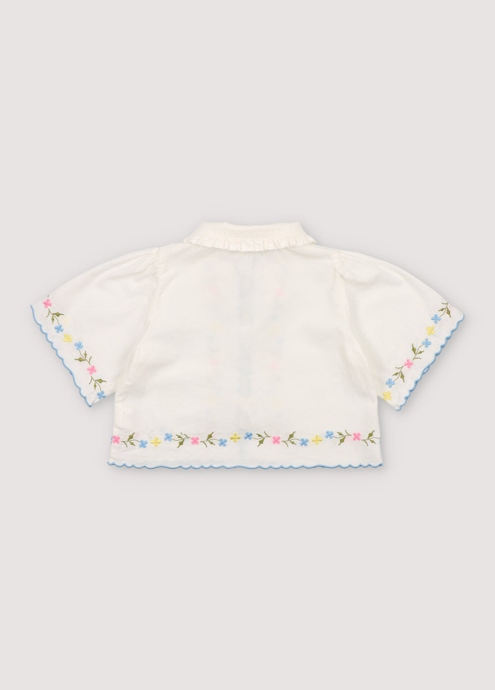 Hermione Blouse in 100% white cotton baize fabric with flower embroidery. Short-sleeved blouse, shirt collar, and central opening with bows, flower embroideries, and powder blue embroidery detail for the finishes.