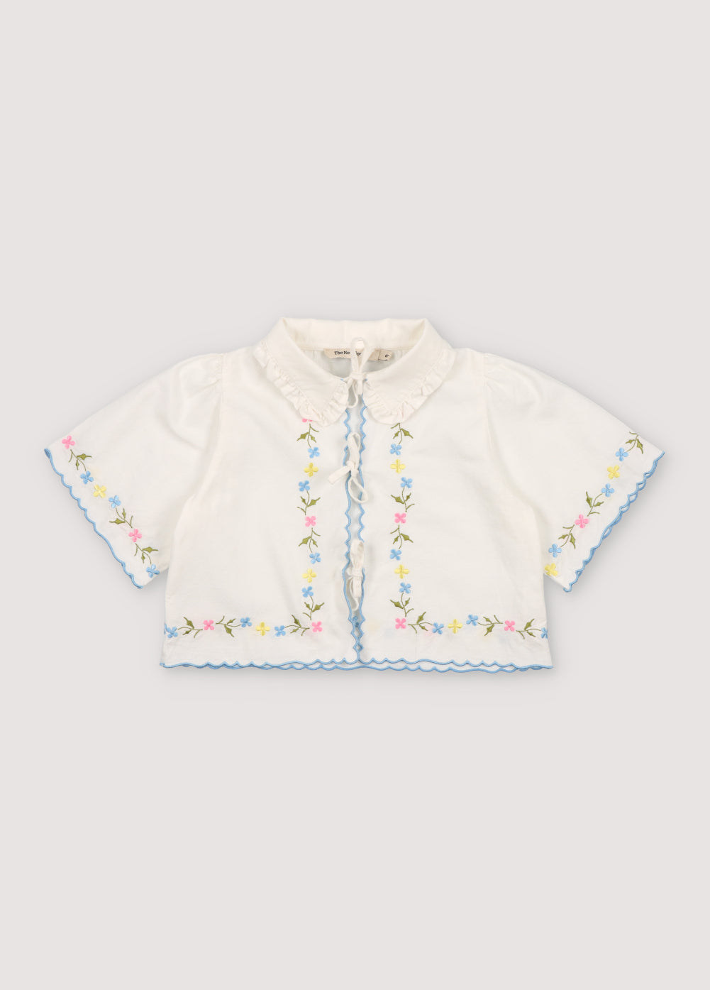 Hermione Blouse in 100% white cotton baize fabric with flower embroidery. Short-sleeved blouse, shirt collar, and central opening with bows, flower embroideries, and powder blue embroidery detail for the finishes.