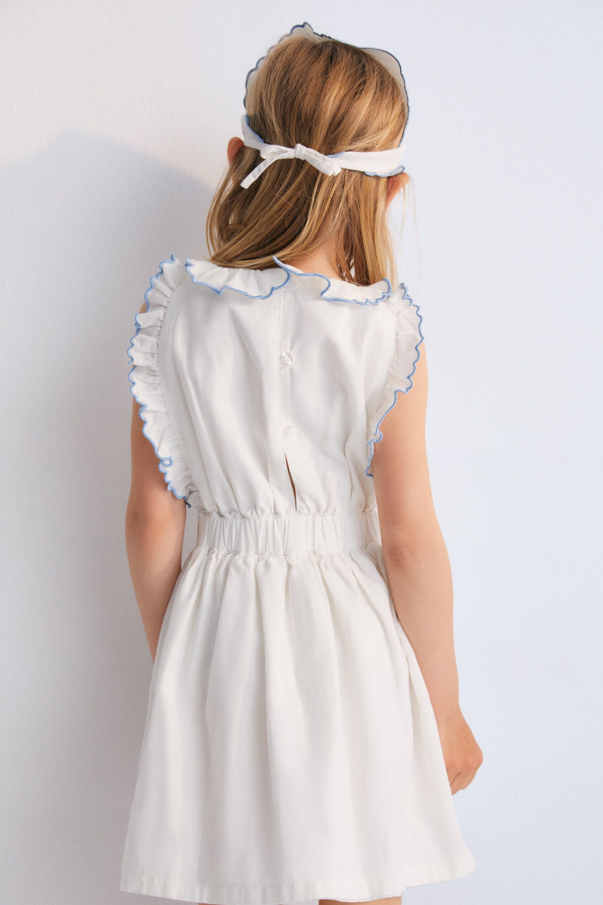 Hermione Dress in 100% white cotton baize fabric with flower embroidery. Dress with gathered neckline, ruffles, and elastic waistband with flower embroidery, and final embroidery detail in powder blue. Spring Summer Collection. The New Society.