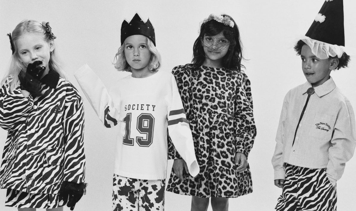 Four children modeling animal-print and zebra-pattern clothing in a black-and-white studio photo. One child wears a zebra-print jacket and pants, another wears a white long-sleeve top with 'SOCIETY 19' and patterned pants, a third wears a leopard-print dress, and a fourth wears a light top with a zebra-print skirt and a pointed hat.
