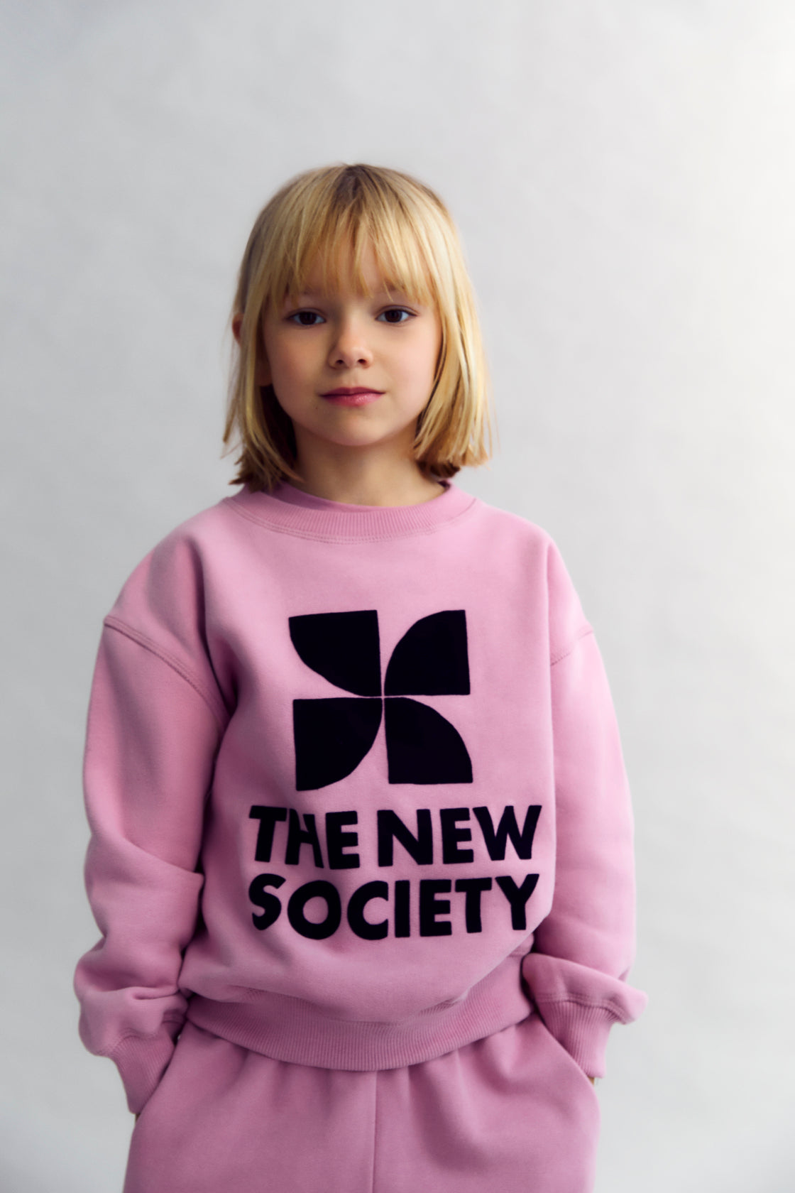 Child wearing a pink sweatshirt with 'The New Society' text and black logo on a plain background