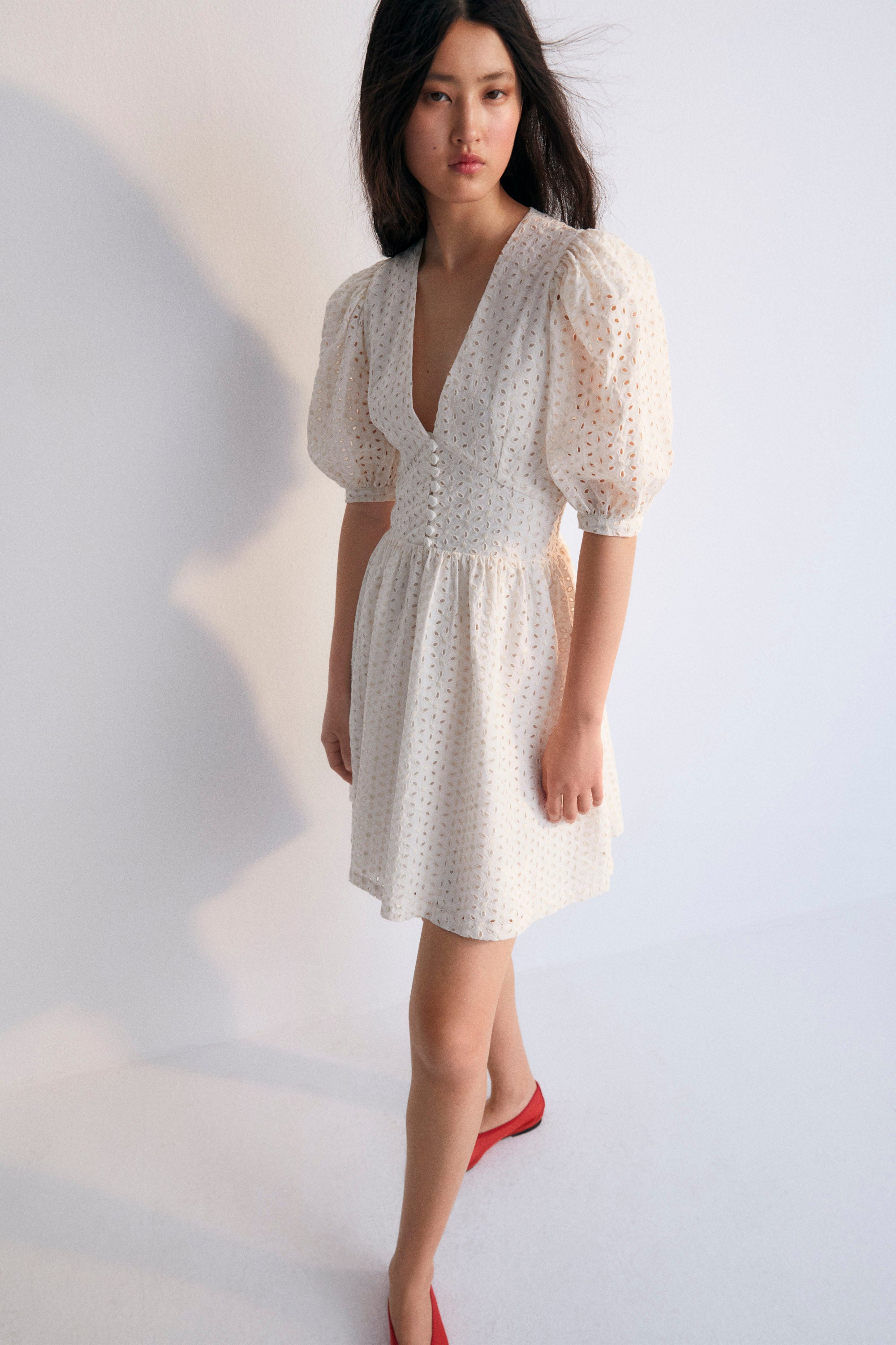 Nemesis Woman Dress in 100% cotton embroidered poplin. It features a V-neck, long billowy sleeves, and a fitted waist. It closes at the front with buttons covered in the same fabric.