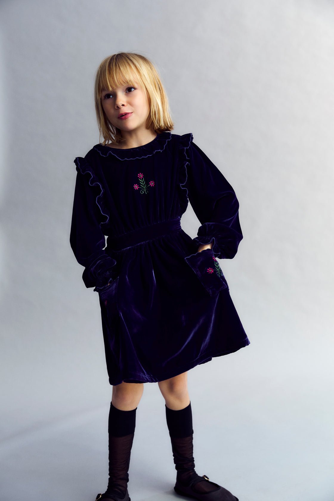 Vestido de velour con cintura entallada, detalle de bordado en el pecho y dos bolsillos bordados en la falda. Está rematado con volantes en los puños y se abre en la espalda con detalles de volantes. El vestido tiene forro de algodón a tono.