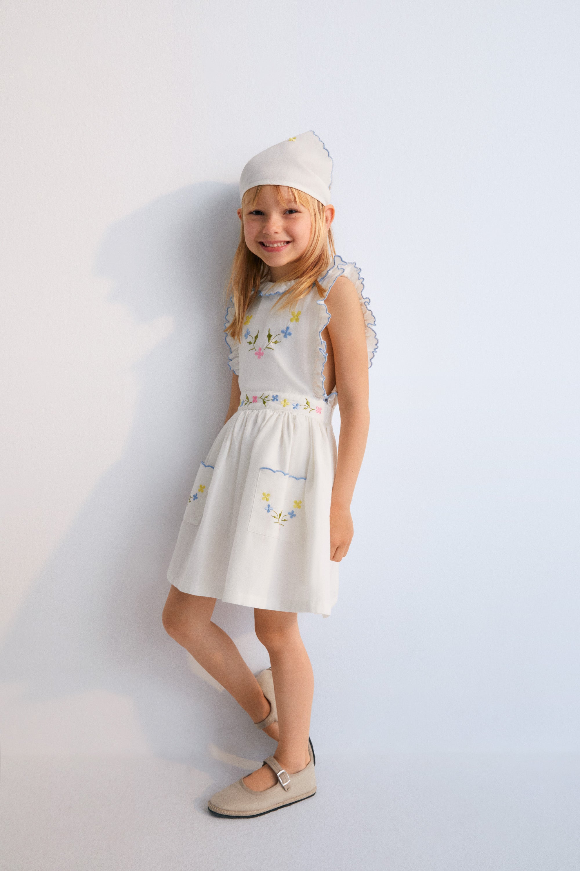 Hermione Dress in 100% white cotton baize fabric with flower embroidery. Dress with gathered neckline, ruffles, and elastic waistband with flower embroidery, and final embroidery detail in powder blue. Spring Summer Collection. The New Society.