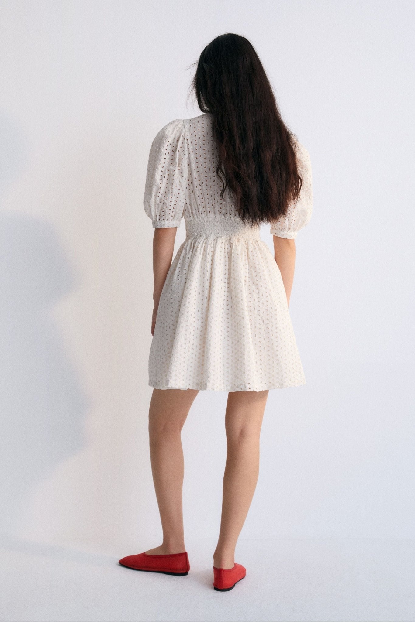 Nemesis Woman Dress in 100% cotton embroidered poplin. It features a V-neck, long billowy sleeves, and a fitted waist. It closes at the front with buttons covered in the same fabric.