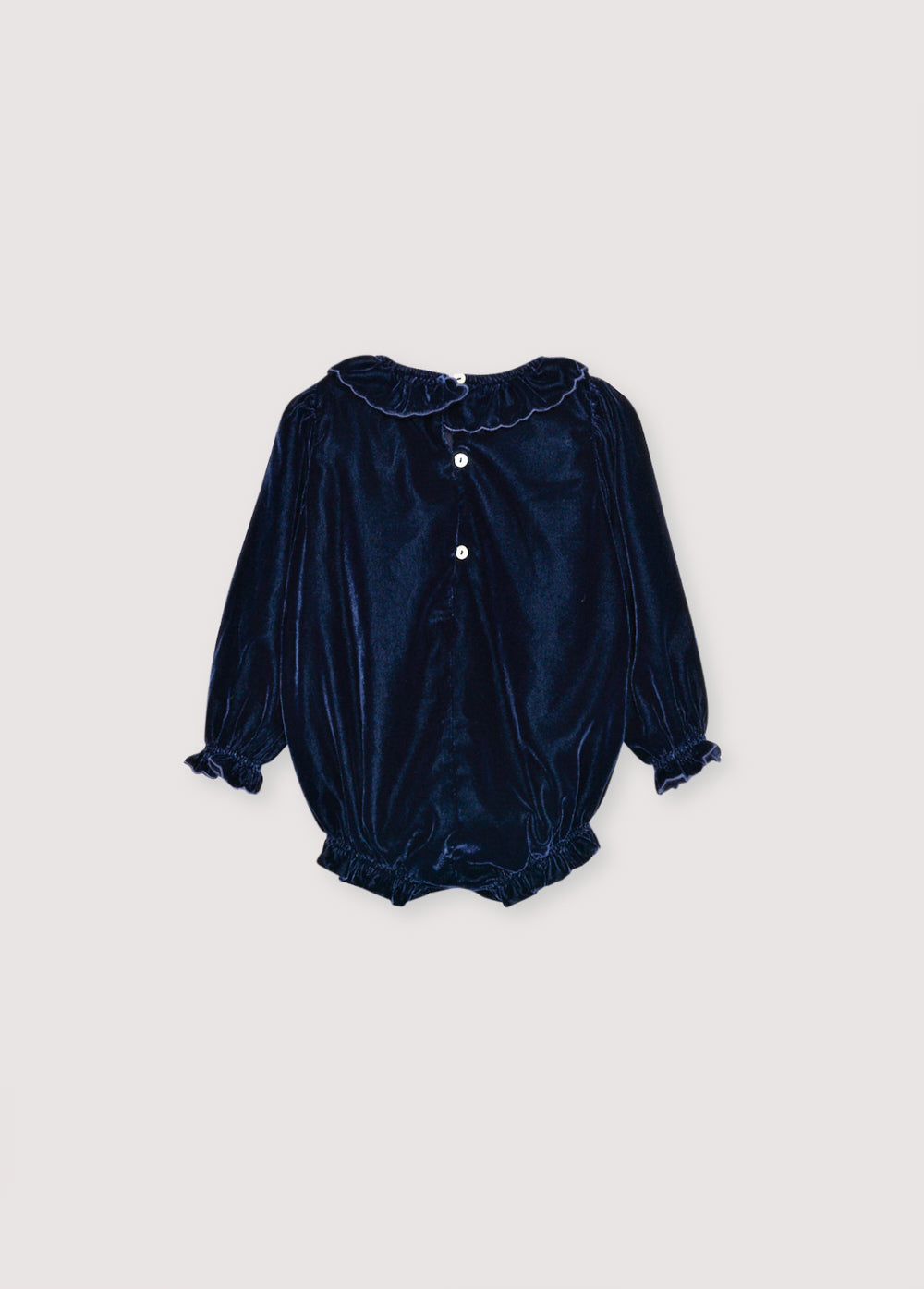 Baby romper in velour with contrasting embroidery detail on the chest. It features a matching fabric collar and a ruffled hem. It opens at the bottom with tone-on-tone snaps and at the back with buttons. it has cotton lining in tone.