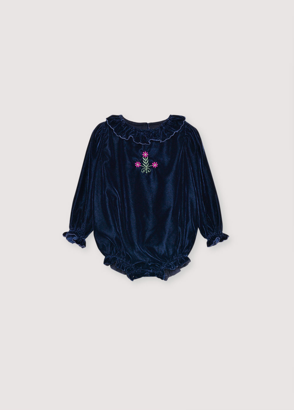 Baby romper in velour with contrasting embroidery detail on the chest. It features a matching fabric collar and a ruffled hem. It opens at the bottom with tone-on-tone snaps and at the back with buttons. it has cotton lining in tone.