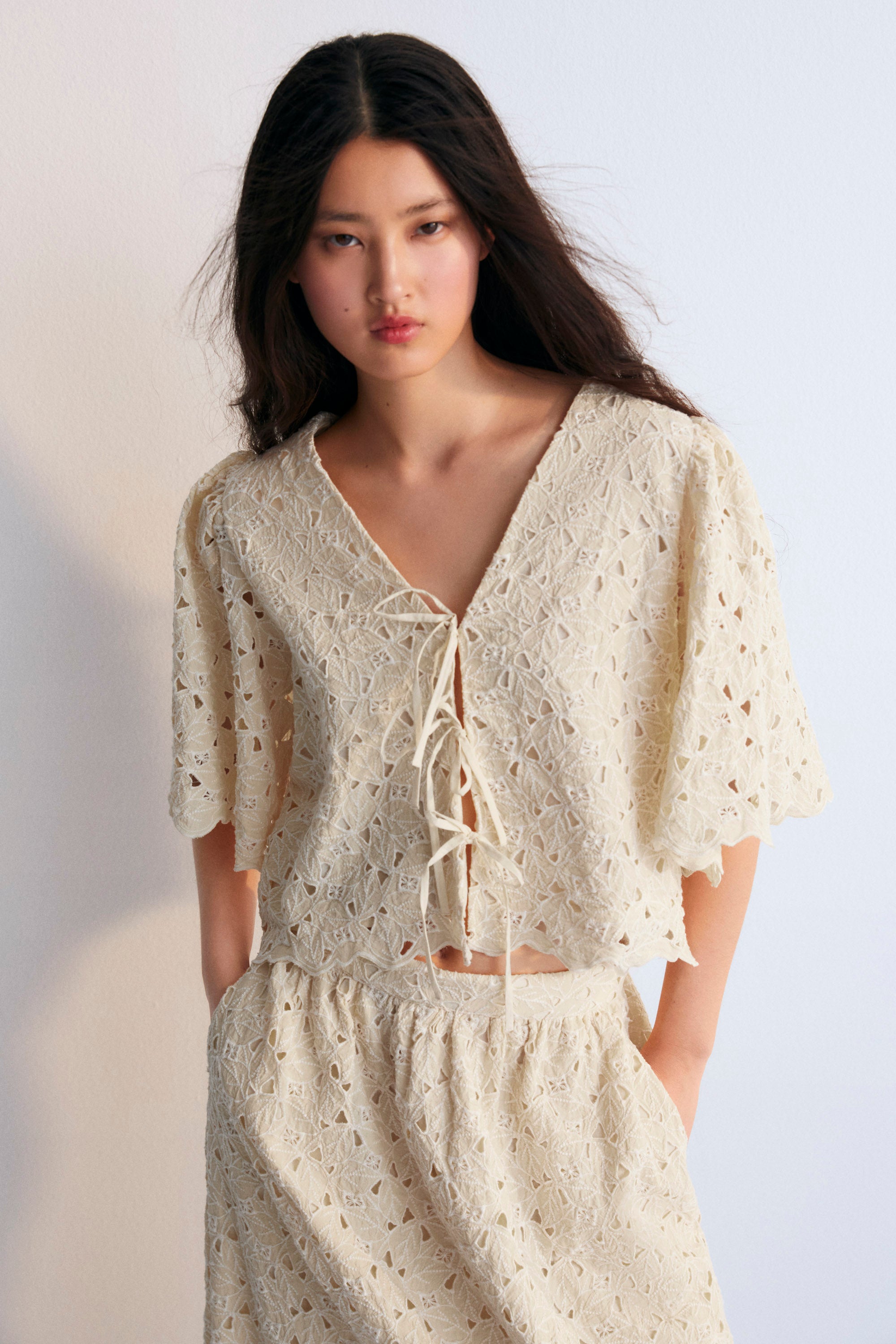 A complete outfit from The New Society's Spring Summer Collection, featuring the Atenea Woman Blouse in embroidered macadamia-colored cotton blend fabric, with gathered sleeves, a V-neckline, and a front closure tied with two bows. Paired with a matching top and pants in the same macadamia tone, creating a cohesive, elegant, and airy look perfect for warmer seasons.