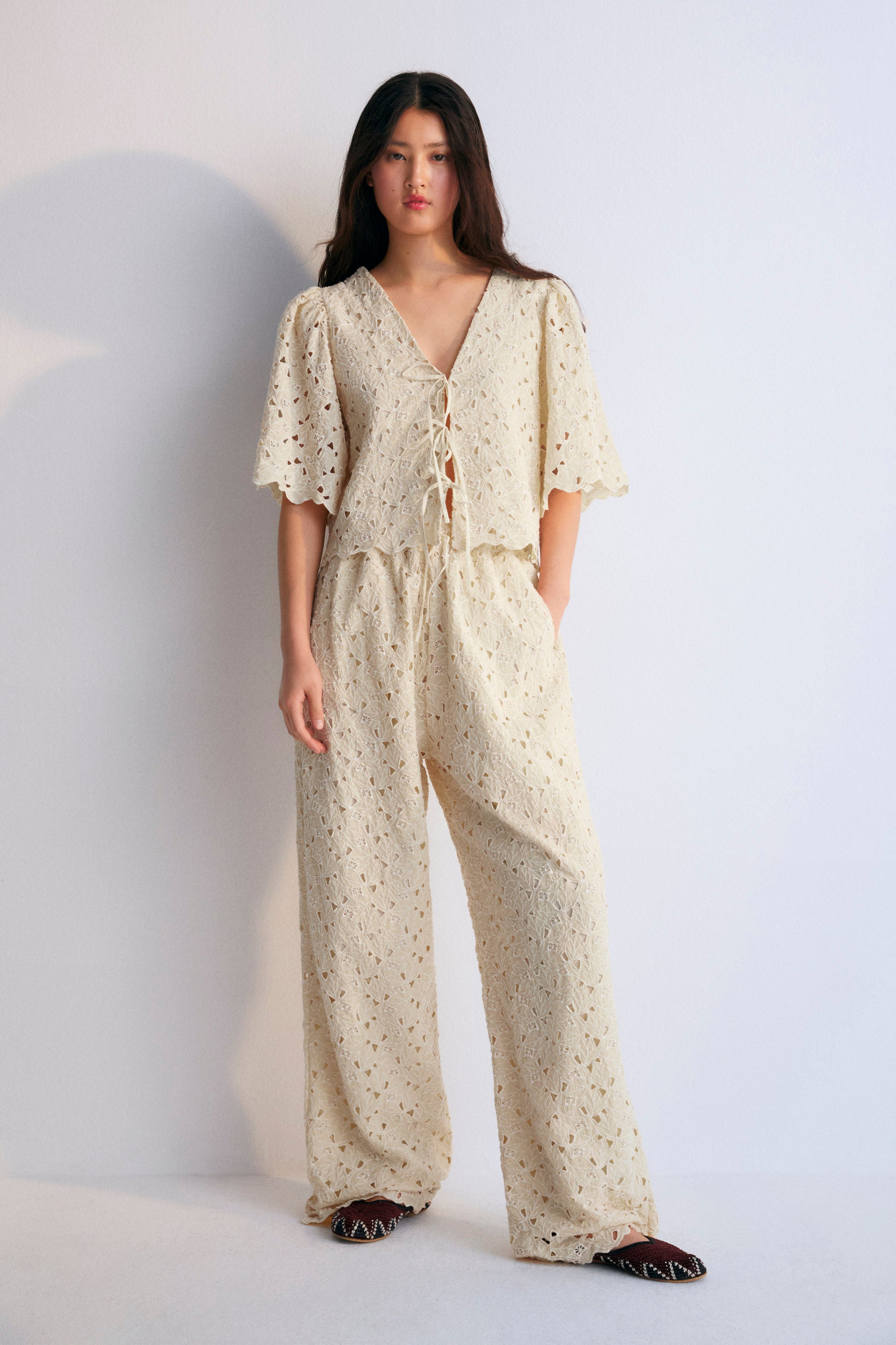 A complete outfit from The New Society's Spring Summer Collection, featuring the Atenea Woman Blouse in embroidered macadamia-colored cotton blend fabric, with gathered sleeves, a V-neckline, and a front closure tied with two bows. Paired with a matching top and pants in the same macadamia tone, creating a cohesive, elegant, and airy look perfect for warmer seasons.