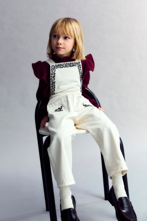 Girl wearing The New Society Tamara Overall in Moonlight White — embroidered denim jumpsuit with adjustable shoulder straps and elasticized back waistband.