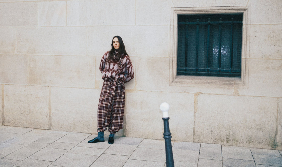 A woman stands against a beige stone wall wearing a long-sleeve, high-waisted plaid jumpsuit in brown, red, and black. She is standing next to a dark barred window on a stone pavement.