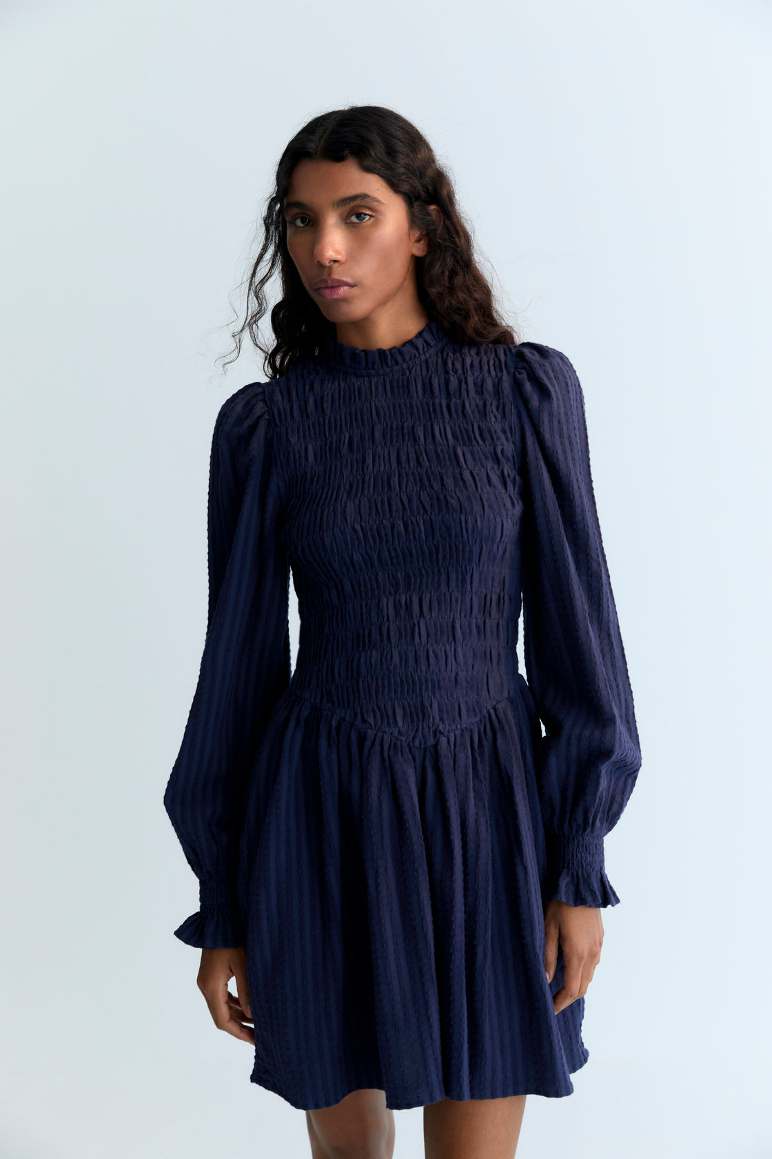 Short seersucker dress with a smocked bodice and V-shaped finish. It features puffed sleeves and a round neckline with a ruffle detail.