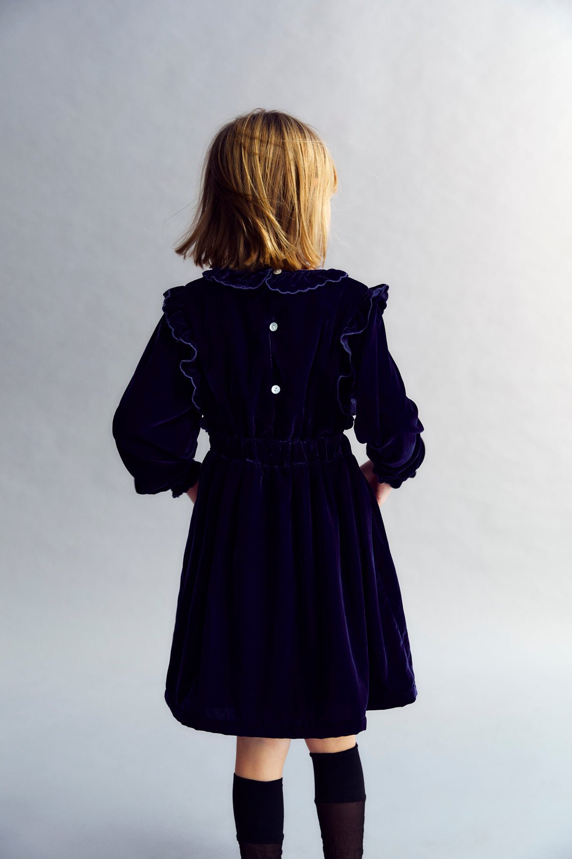 Vestido de velour con cintura entallada, detalle de bordado en el pecho y dos bolsillos bordados en la falda. Está rematado con volantes en los puños y se abre en la espalda con detalles de volantes.  El vestido tiene forro de algodón a tono. 