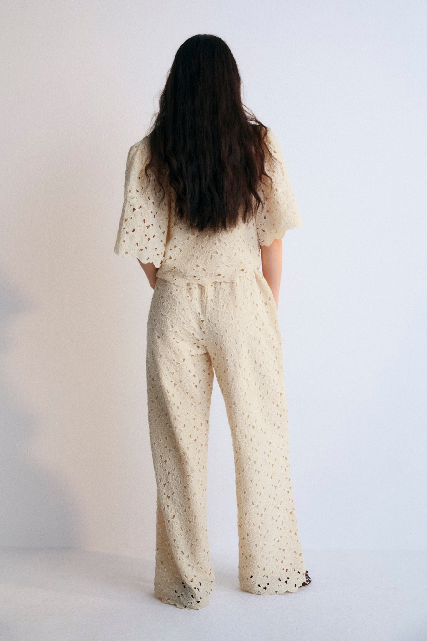 A complete outfit from The New Society's Spring Summer Collection, featuring the Atenea Woman Blouse in embroidered macadamia-colored cotton blend fabric, with gathered sleeves, a V-neckline, and a front closure tied with two bows. Paired with a matching top and pants in the same macadamia tone, creating a cohesive, elegant, and airy look perfect for warmer seasons.