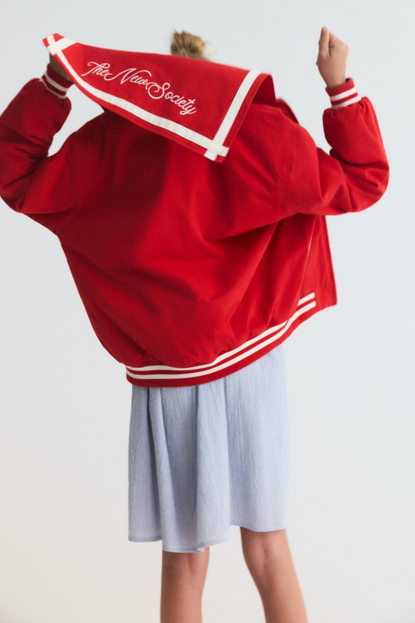 Chaqueta estilo bomber en un rojo vibrante, con un exclusivo cuello marinero, logotipo de la Sociedad bordado en la espalda y rayas en contraste. 