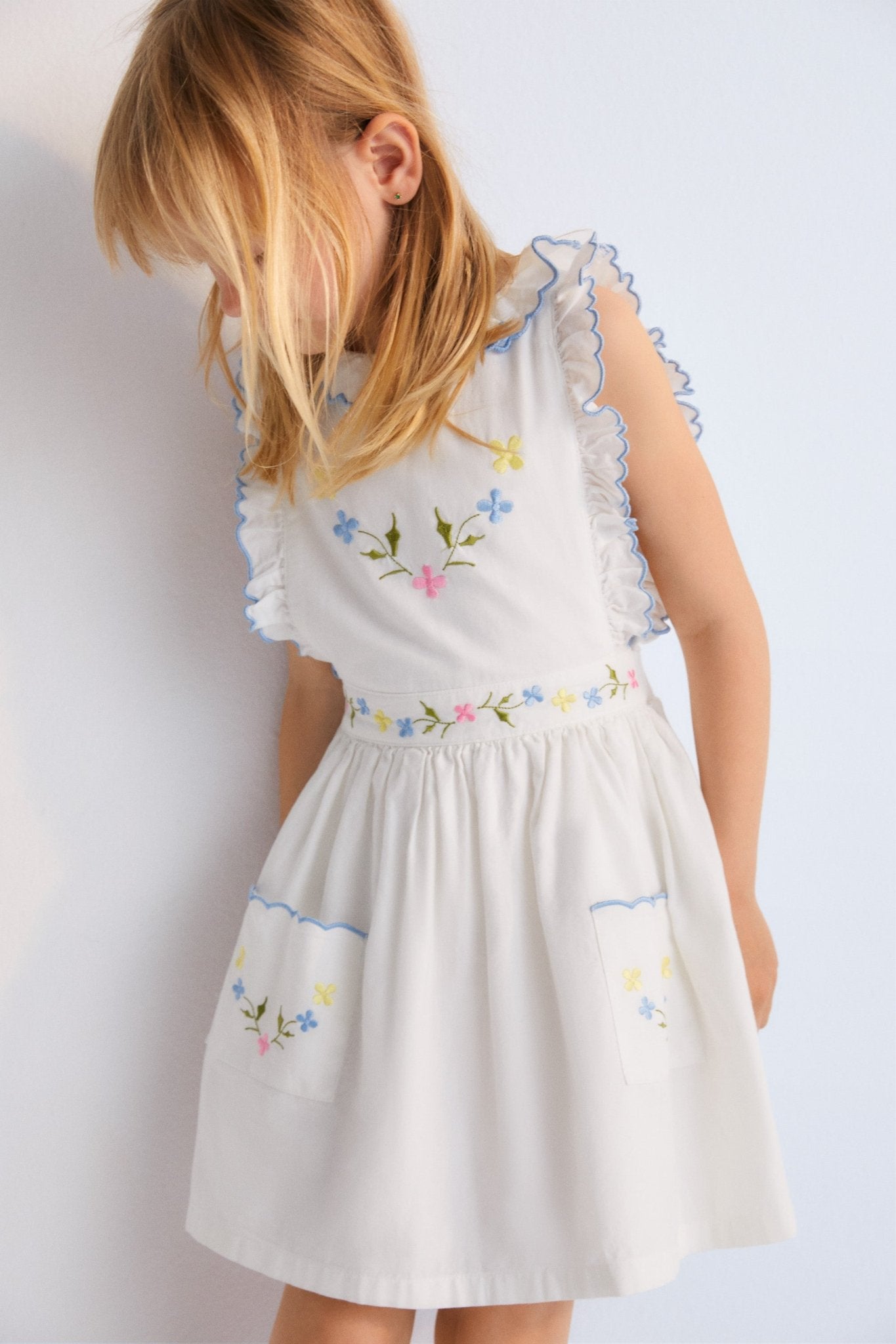 Hermione Dress in 100% white cotton baize fabric with flower embroidery. Dress with gathered neckline, ruffles, and elastic waistband with flower embroidery, and final embroidery detail in powder blue. Spring Summer Collection. The New Society. 
