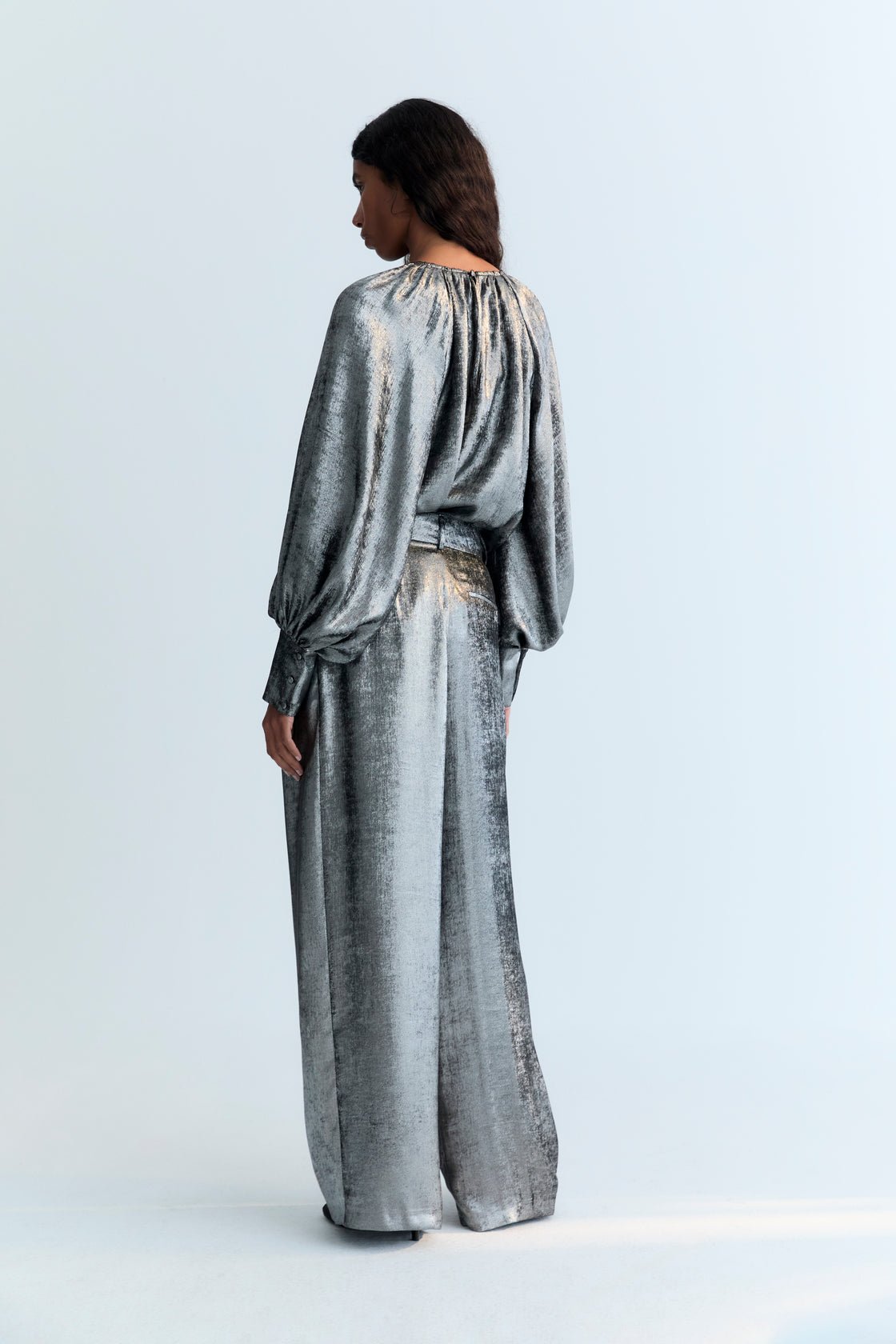 Pantalón recto en tejido brillo plateado con detalle de pliegues delanteros, bolsillos y cinturilla cruzada con botón oculto.
